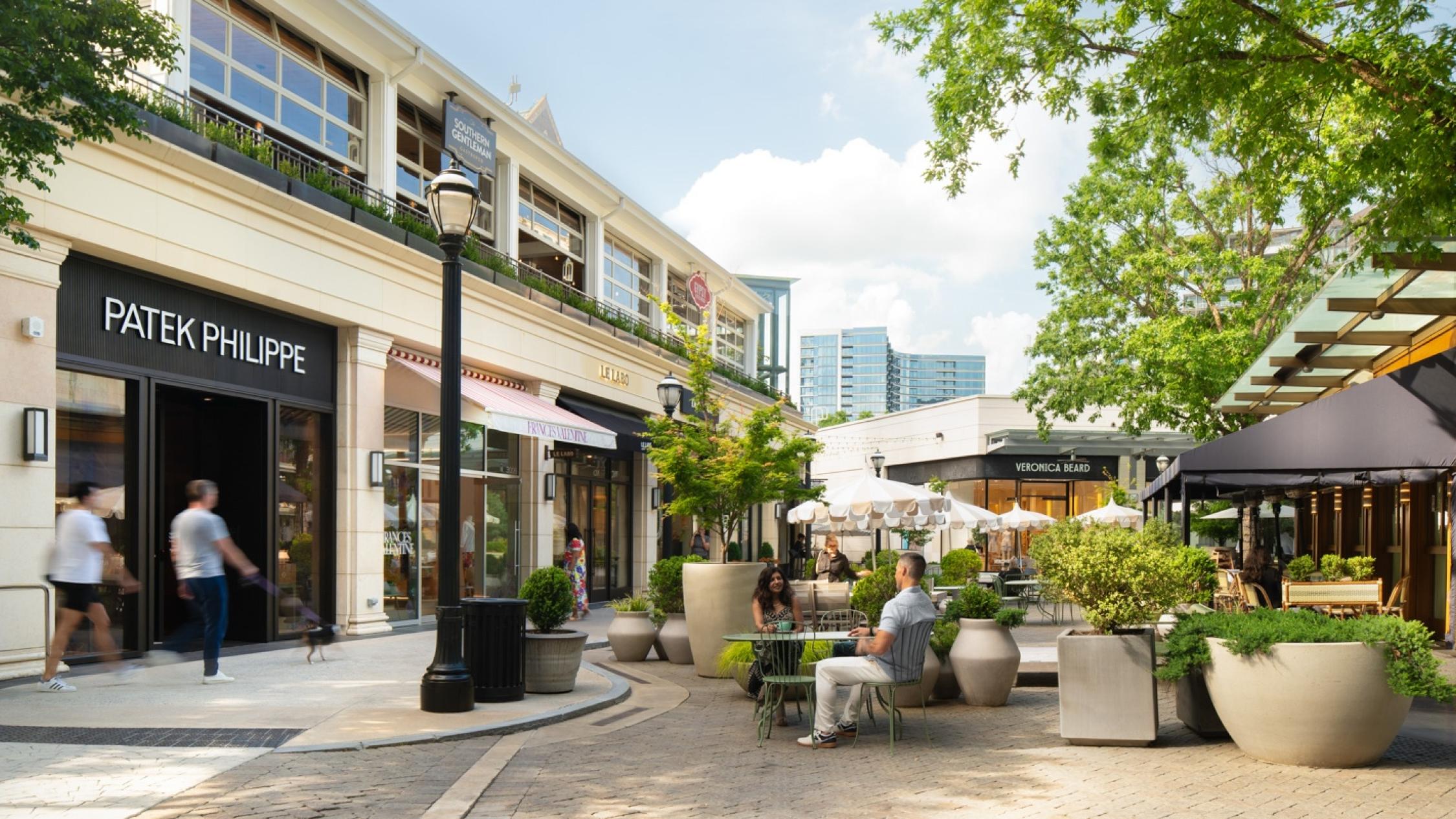 People walking on sidewalks in front of shops, and sitting at tables on a patio, in an outdoor area at Buckhead Village