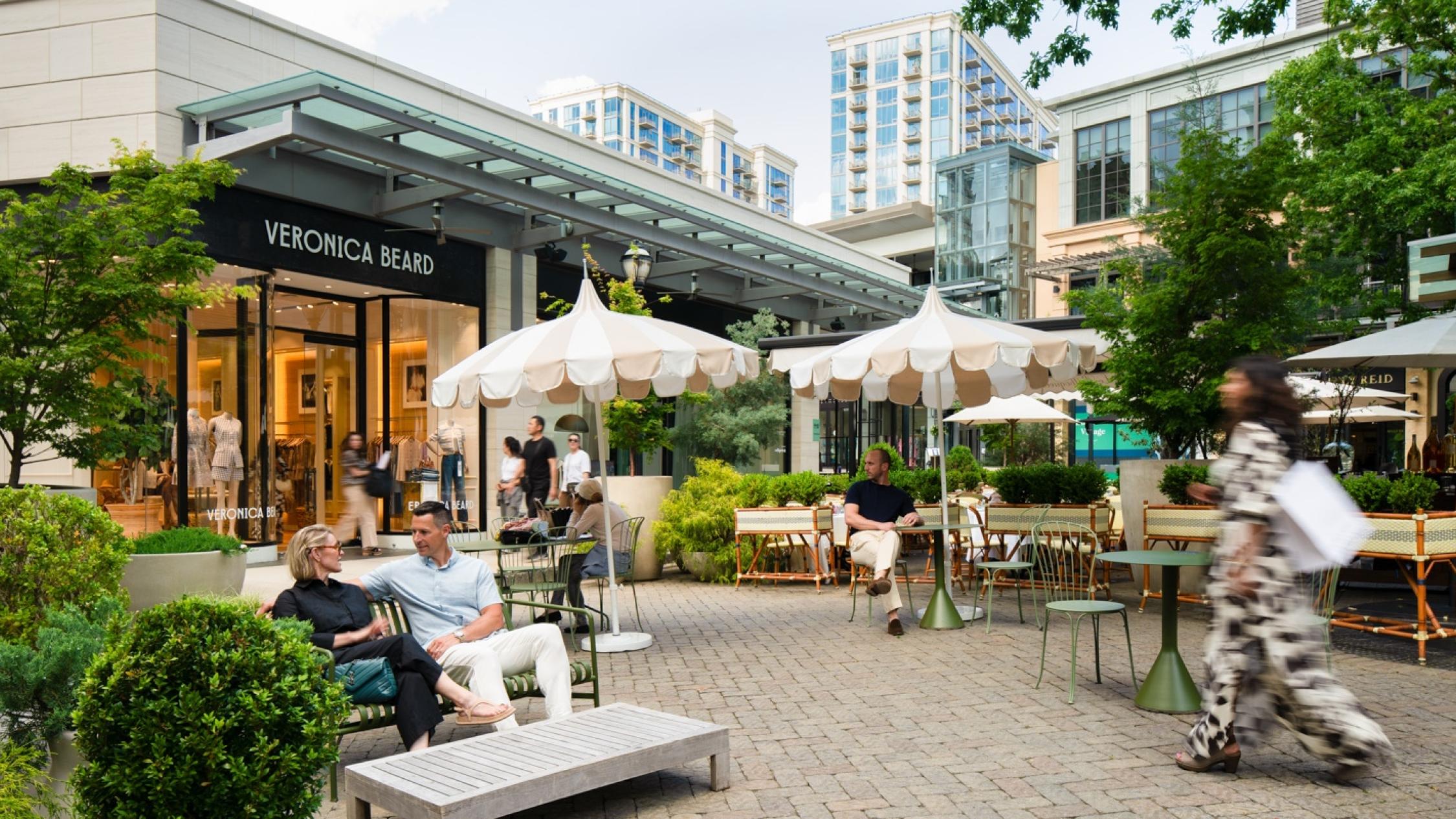 People walking on sidewalks in front of shops, and sitting on a bench and at tables on a patio, in an outdoor area at Buckhead Village