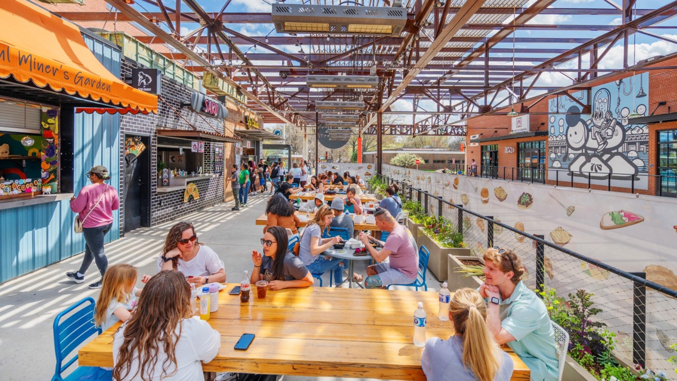 People sitting and eating food at outdoor tables along a sidewalk in front of Camp North End eateries