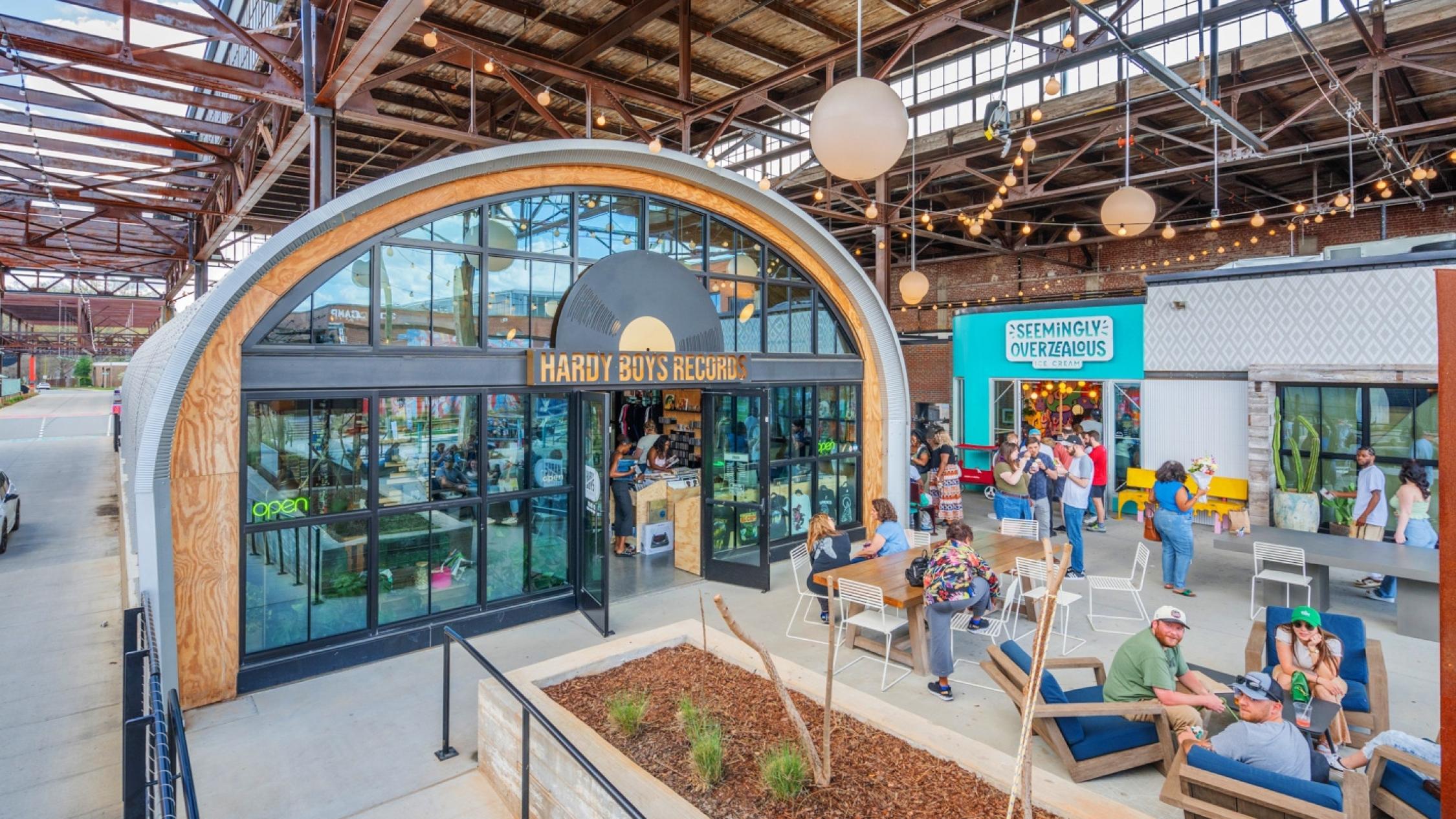 People standing and seated at tables in an outdoor common area in front shops at Camp North End