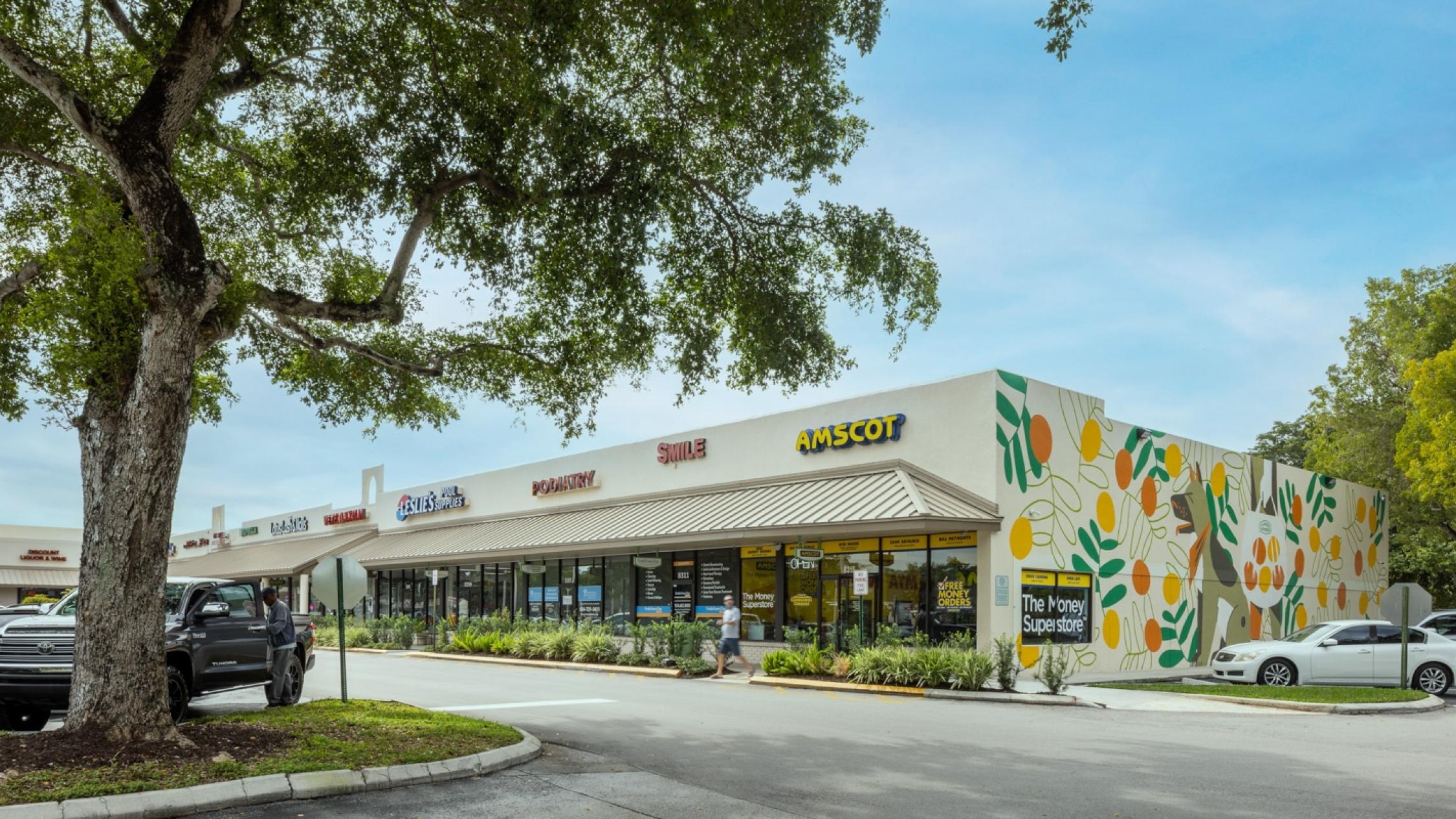 Various store facades with a pedestrian on the sidewalk and cars in the parking lot at Cypress Commons