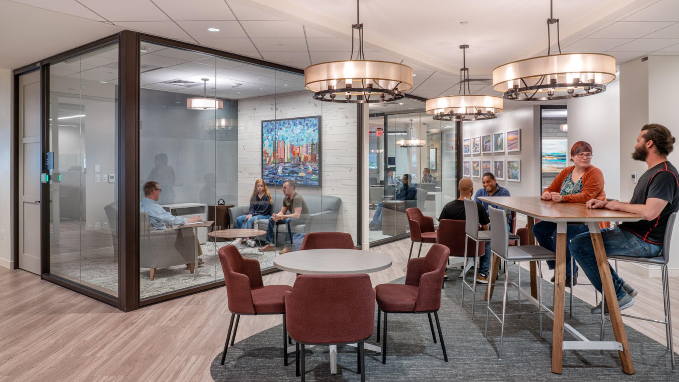 Office interior common area with employees conversing at tables, and a glass-enclosed conference room with people in a meeting