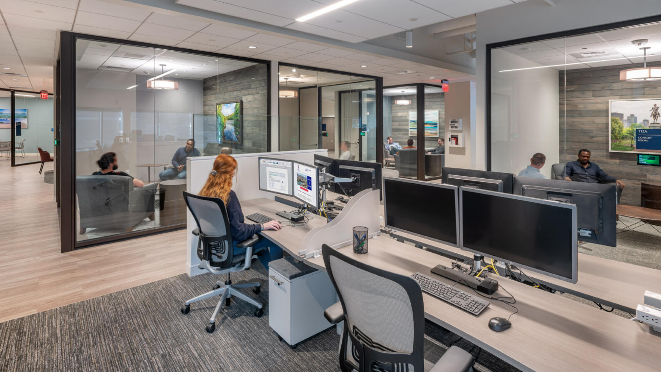 Open floor plan office space with a person working at a desk, and several glass-enclosed conference rooms with people in meetings