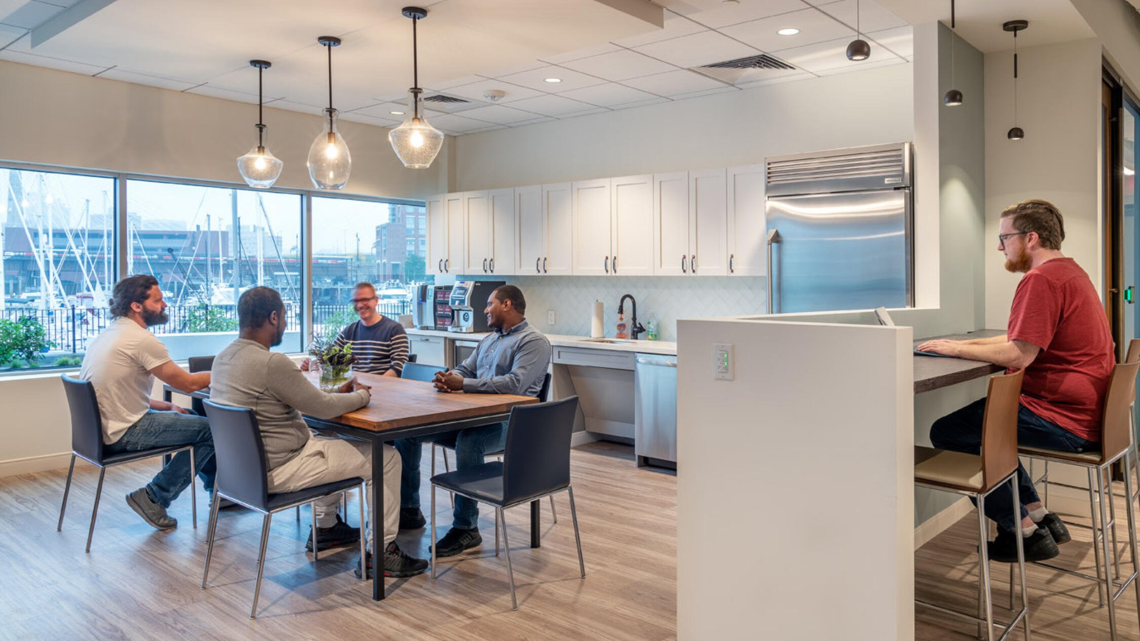 Office break room with employees conversing at a table near a kitchenette, with a view of moored sailboats out the window behind them