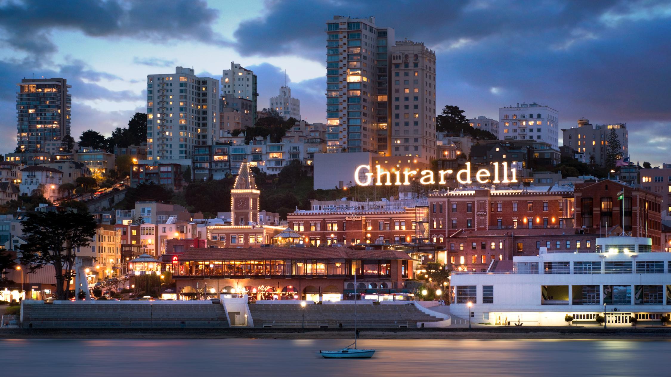 Ghirardelli Square exterior at dusk with bay in foreground and skyline in background