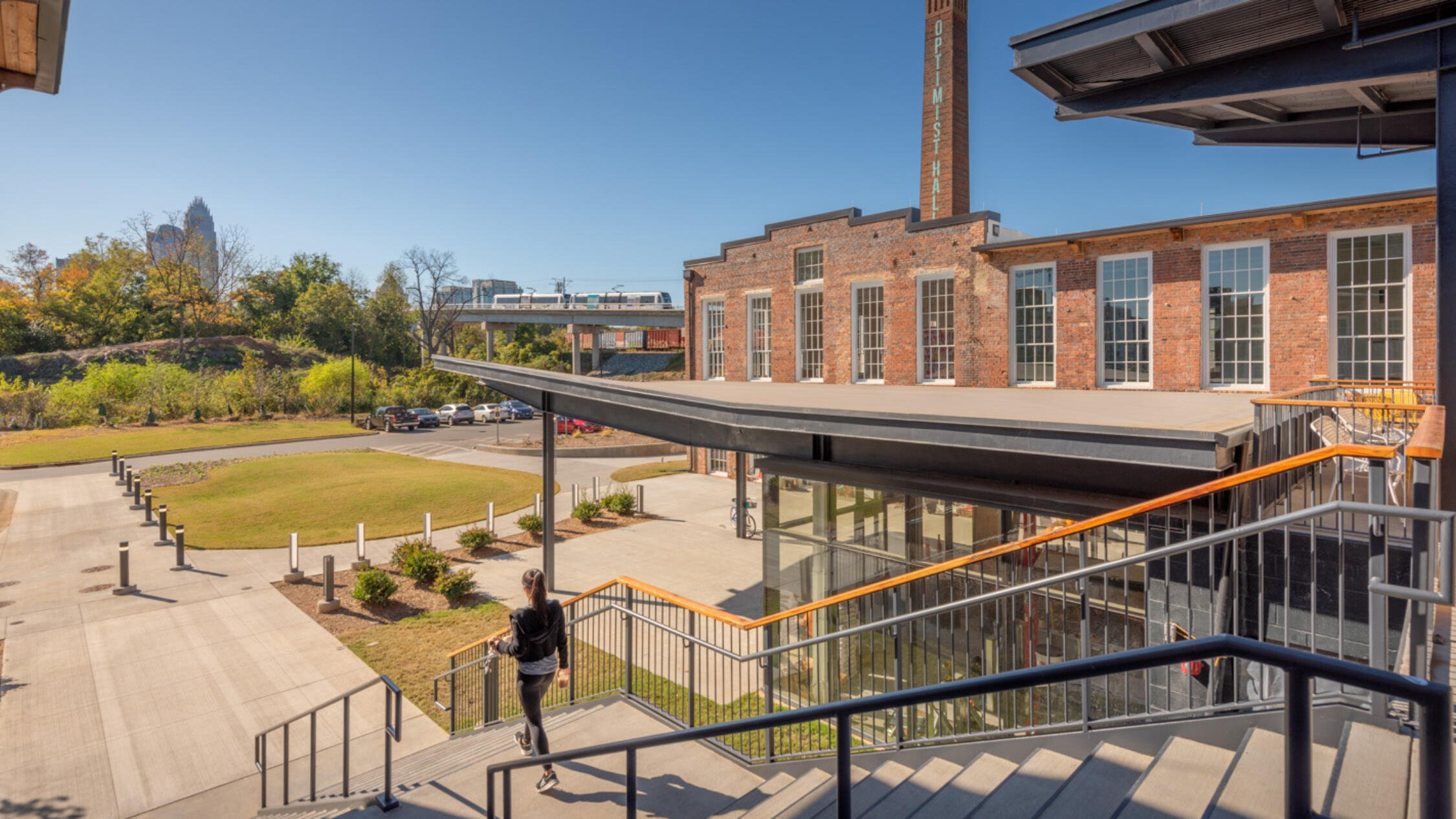 Woman descending outdoor staircase with Optimist Hall building, monument sign, light rail line, and skyline in background