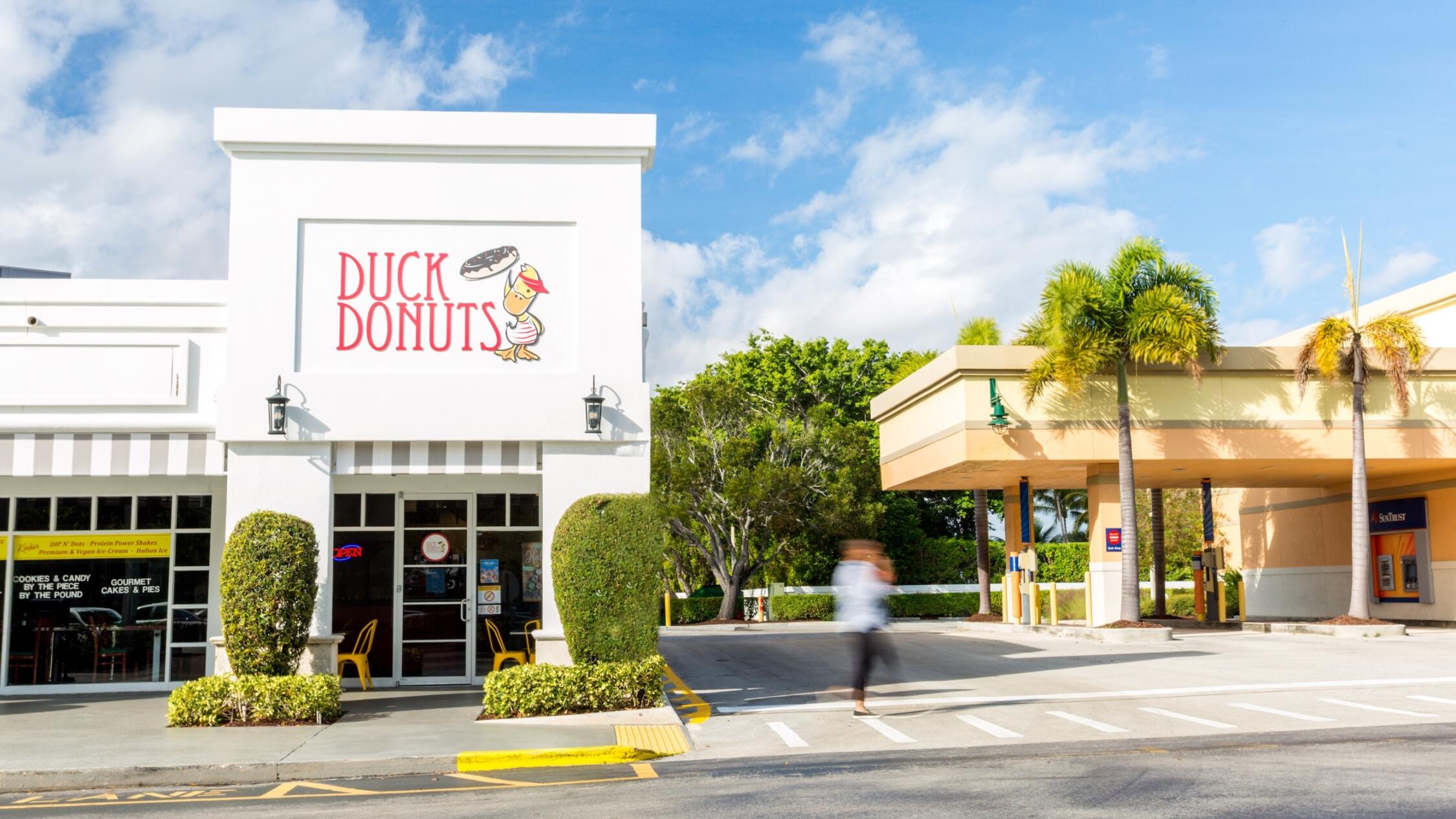 A woman walks by Duck Donuts at Polo Club shops with palm trees and a blue sky.