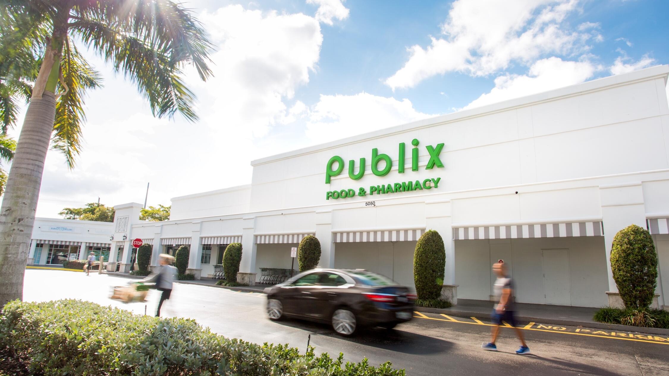 People and a car moving past the Publix in Polo Club Shops with a palm tree on the left and a bright blue sky behind.