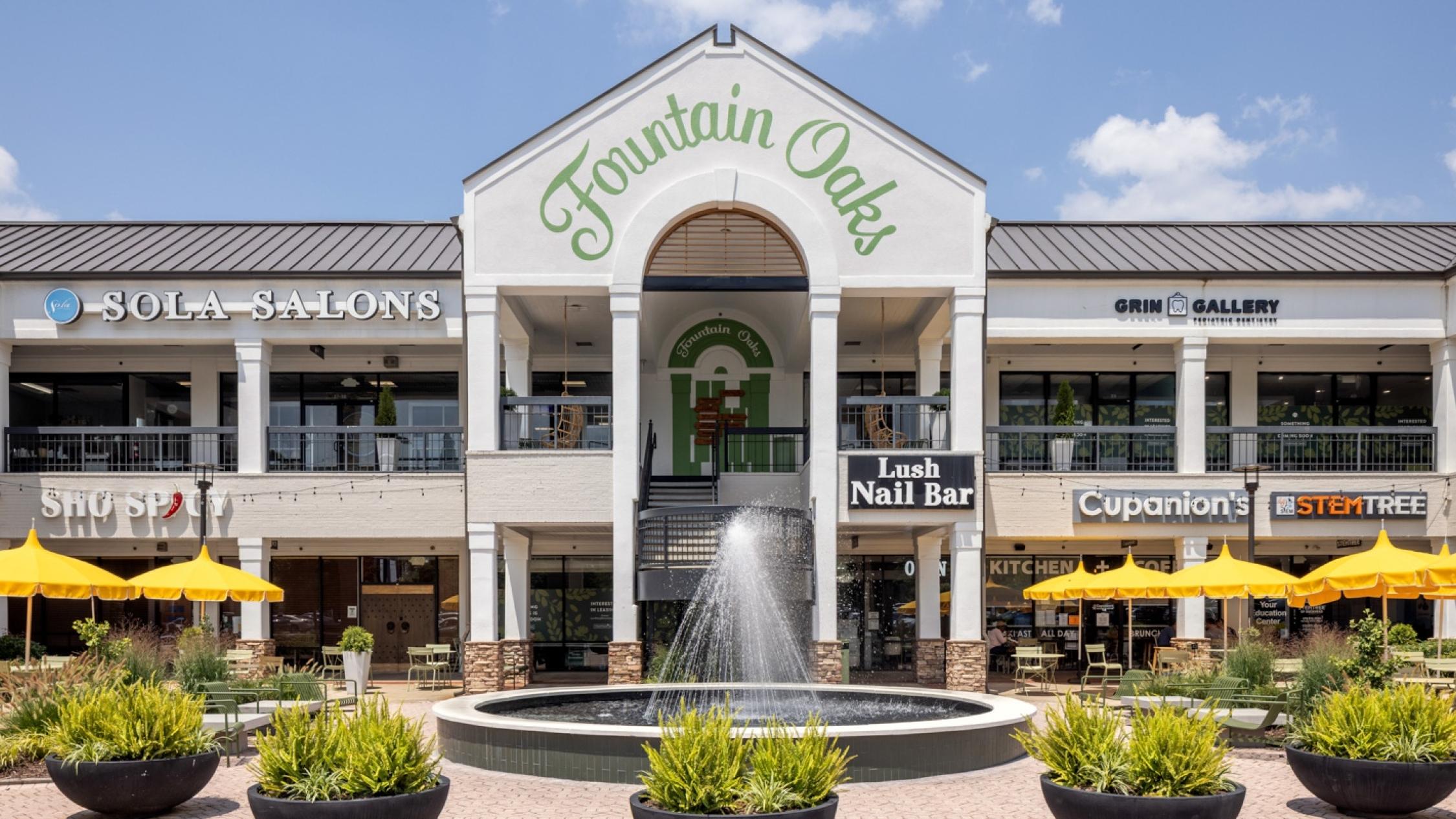 Various store exterior facades in a two-story building with a fountain and tables with umbrellas at Fountain Oaks