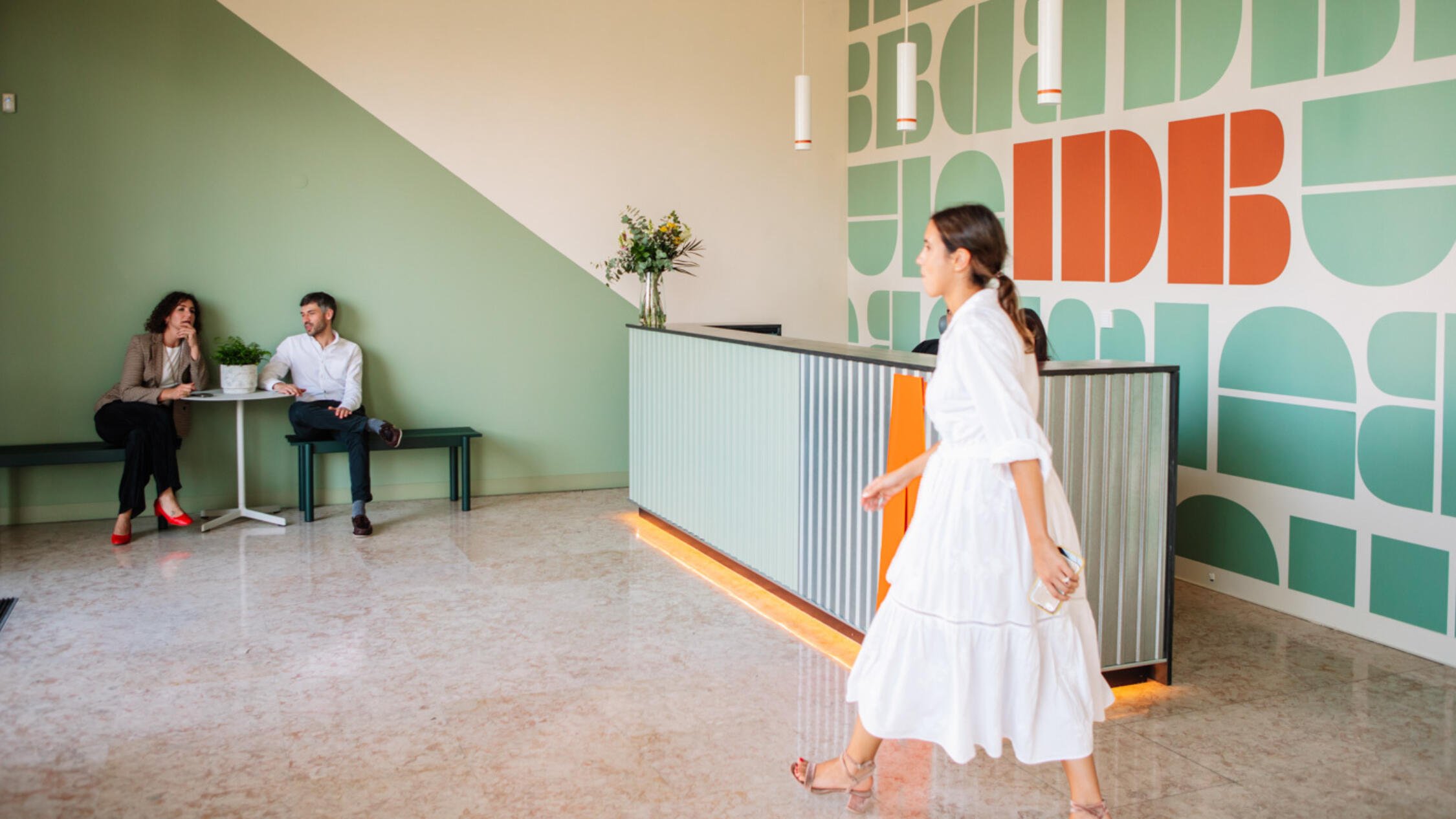 Woman walking past desk in IDB Lisbon's lobby, with man and woman sitting at table in background