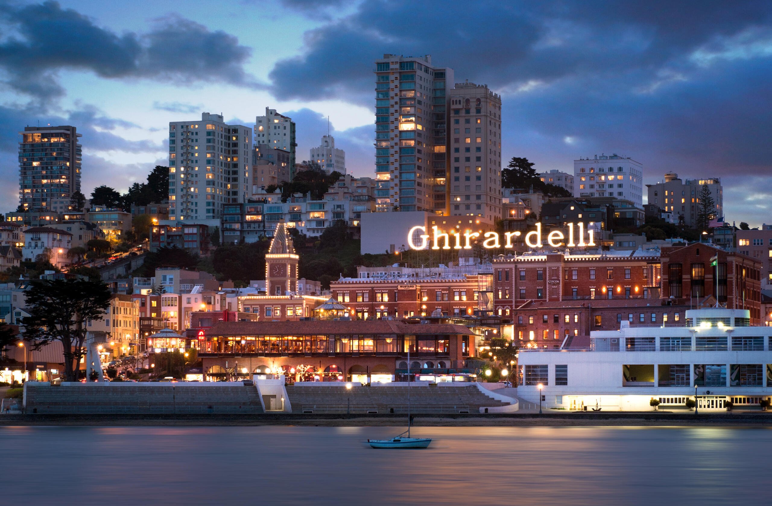 Ghirardelli Square exterior at dusk with bay in foreground and skyline in background