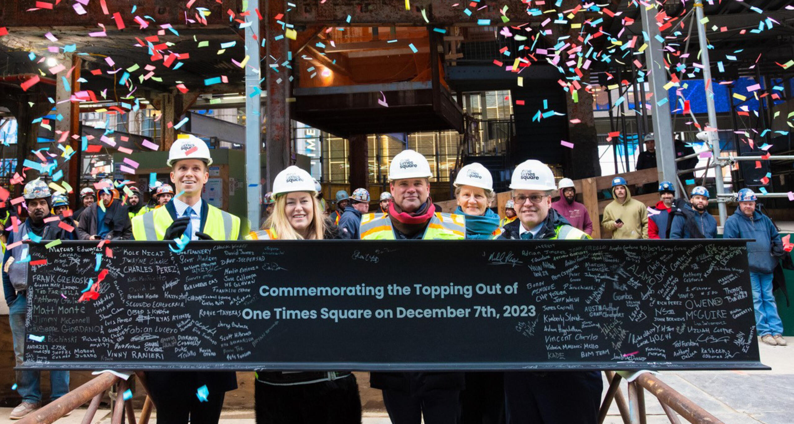 Five people holding a banner that says "commemorating the topping out of One Times Square on December 77th, 2023" with confetti falling in background
