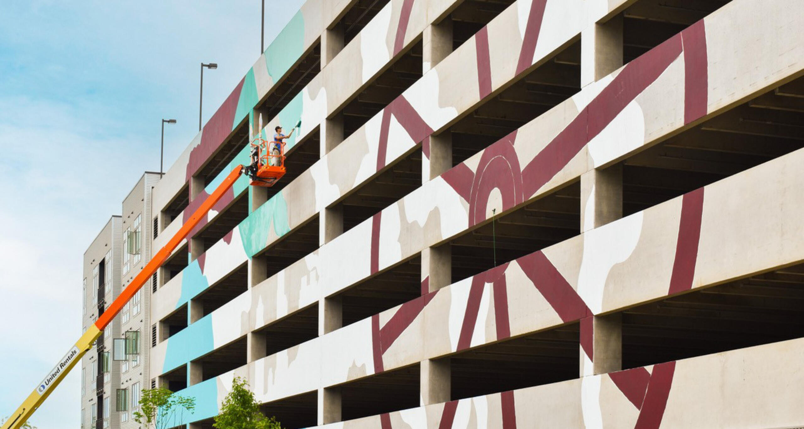 An artist atop a cherry picker crane painting the side of a parking deck at Raleigh Iron Works