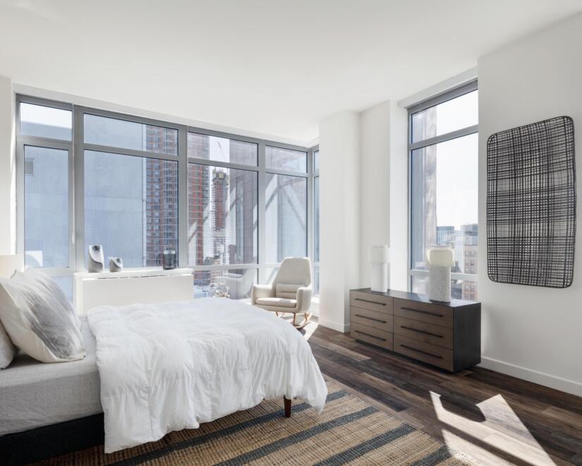 Apartment bedroom with bed, chest of drawers, and chair, with high rise buildings visible outside the windows