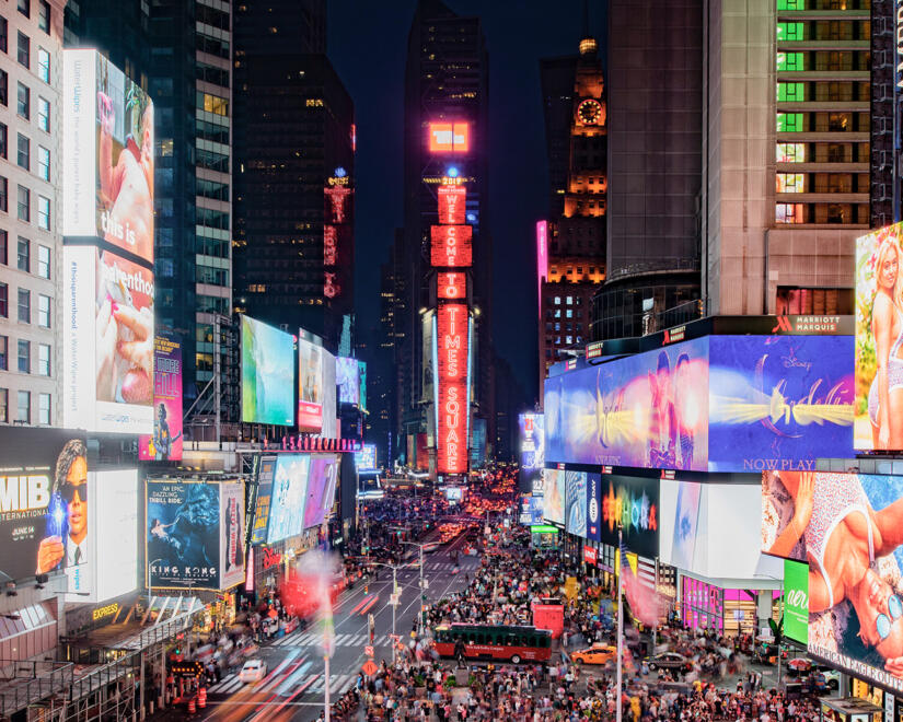 View across One Times Square from mid rise at night with throngs of pedestrians and dozens of illuminated billboards