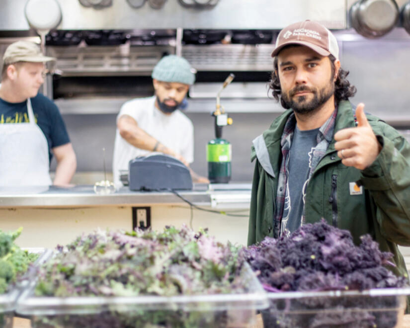 Food Fight GA farmer posing with bins of vegetables in a restaurant kitchen with cooks in the background