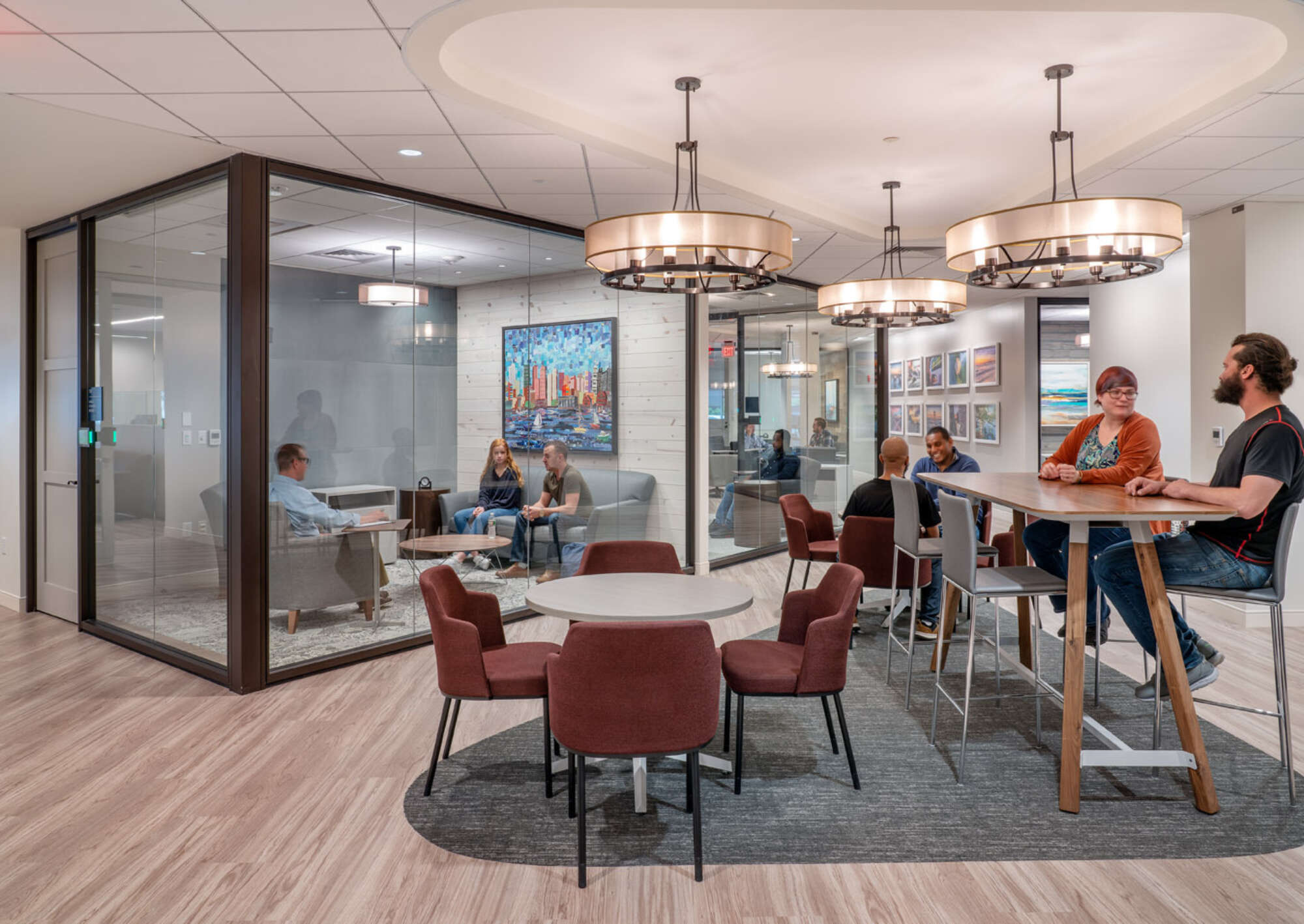 Office interior common area with employees conversing at tables, and a glass-enclosed conference room with people in a meeting