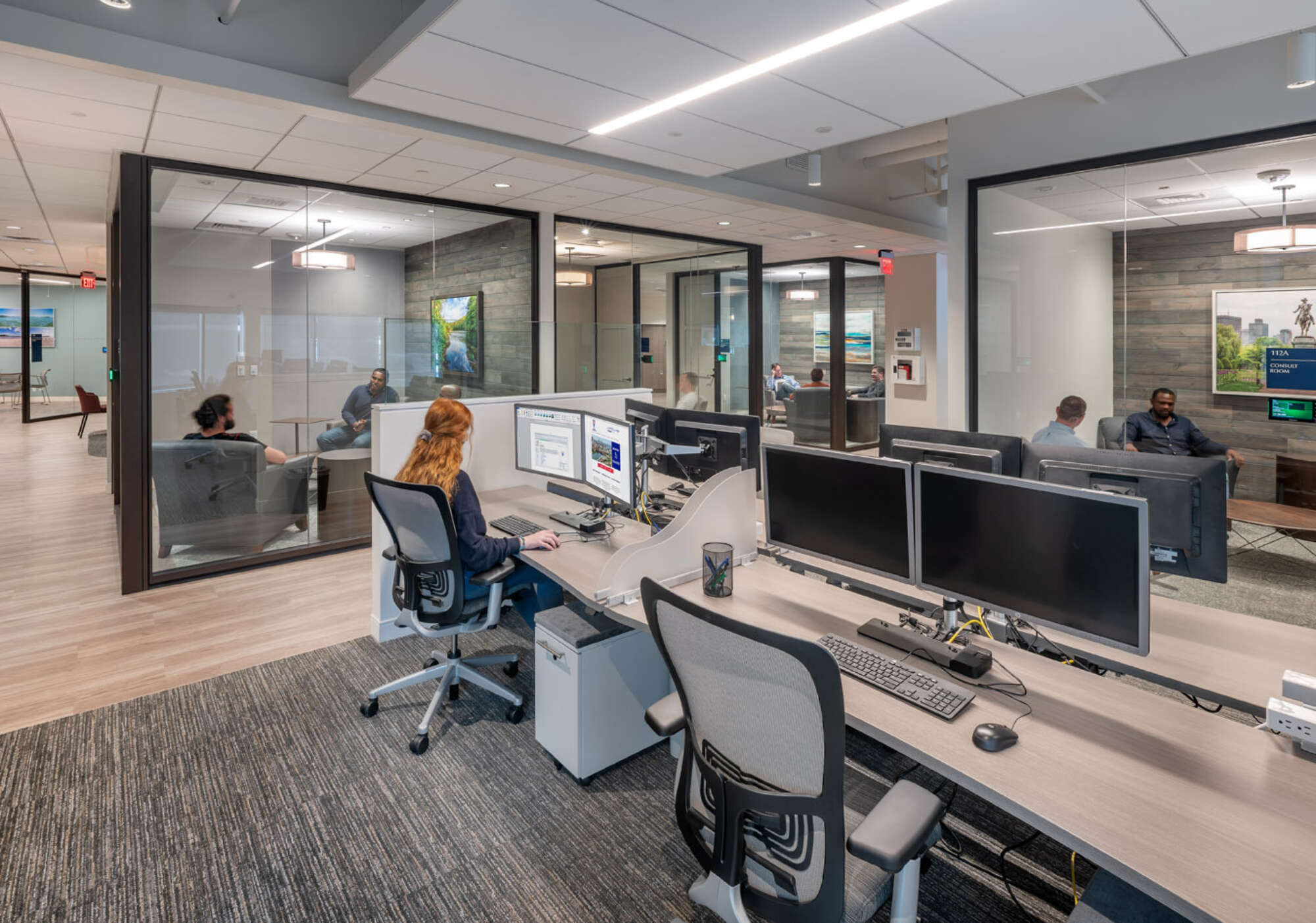 Open floor plan office space with a person working at a desk, and several glass-enclosed conference rooms with people in meetings