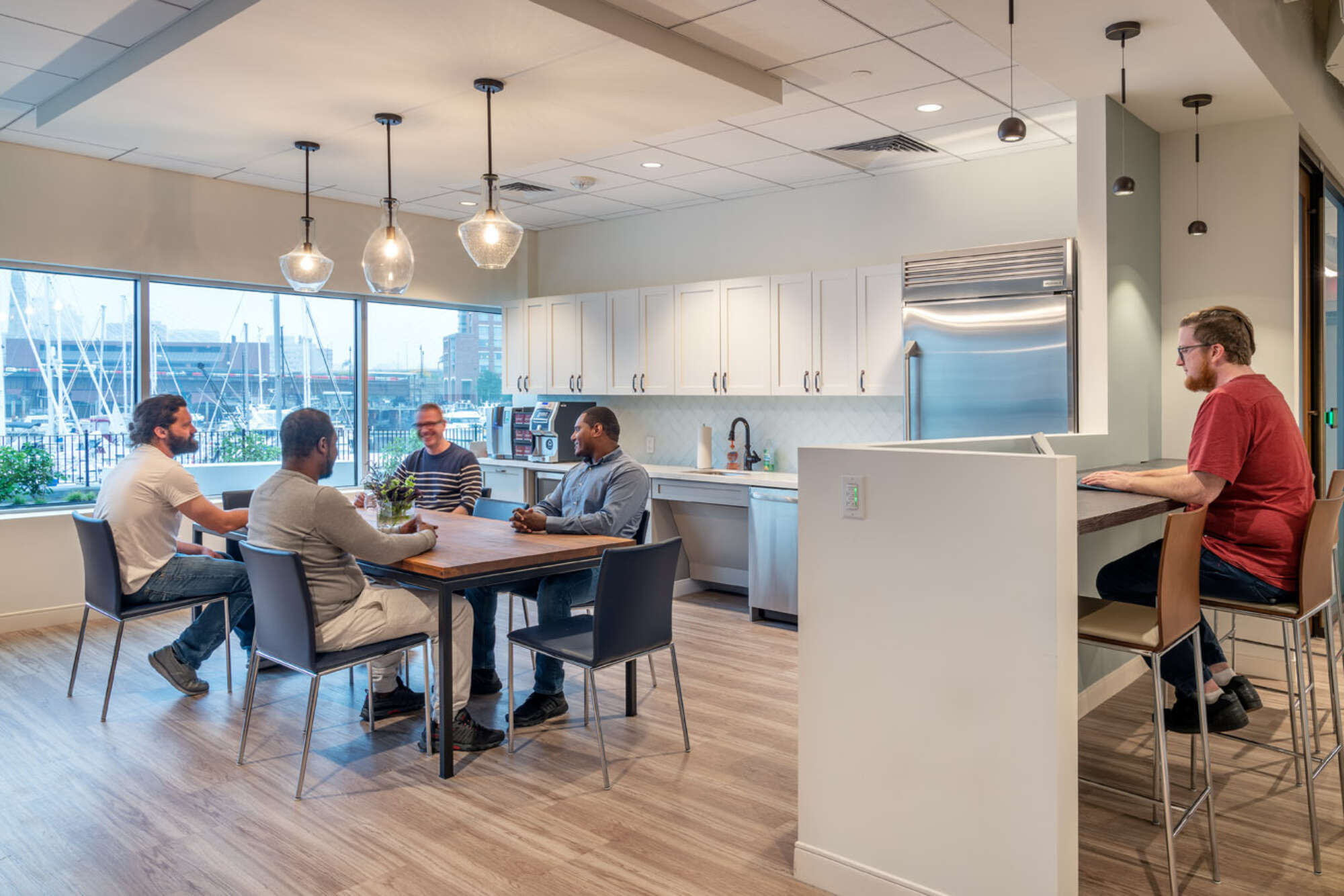 Office break room with employees conversing at a table near a kitchenette, with a view of moored sailboats out the window behind them
