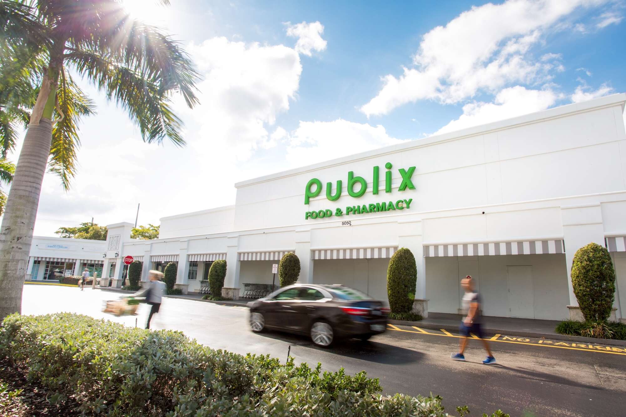 People and a car moving past the Publix in Polo Club Shops with a palm tree on the left and a bright blue sky behind.