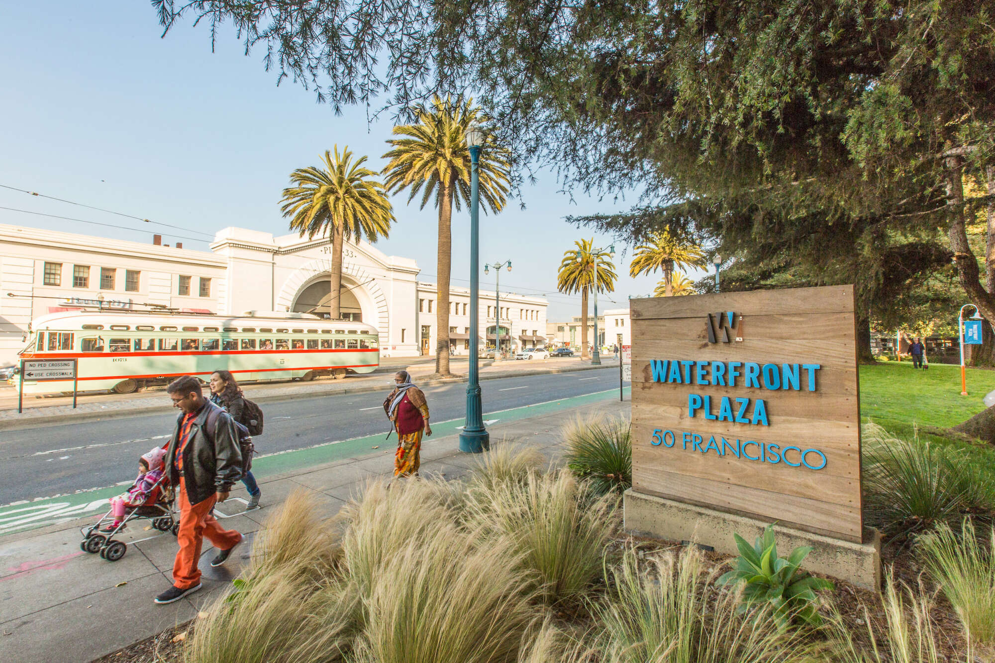 People walk by street front signage of The Waterfront Collection and a San Francisco cable car passes by